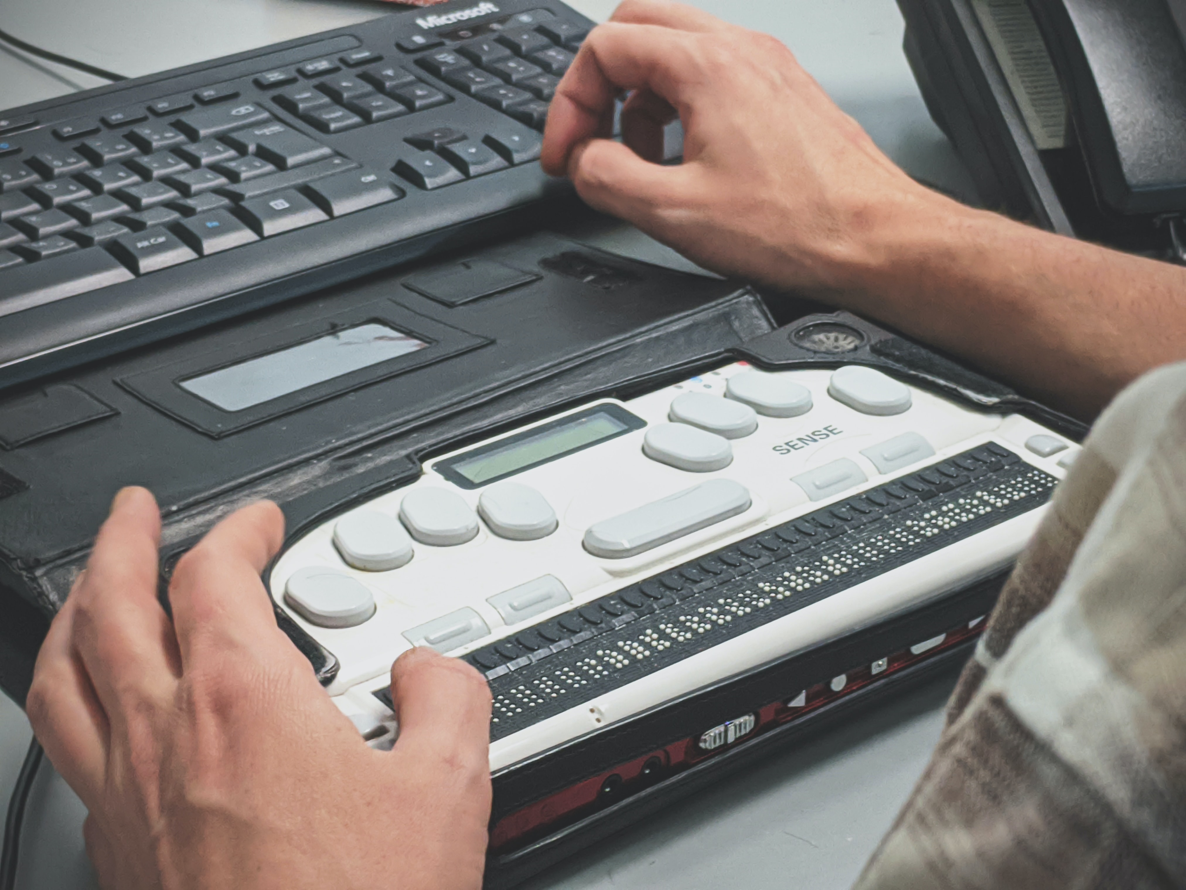 Image of person's hands using HIMS Braille Sense notetaker and Microsoft keyboard.